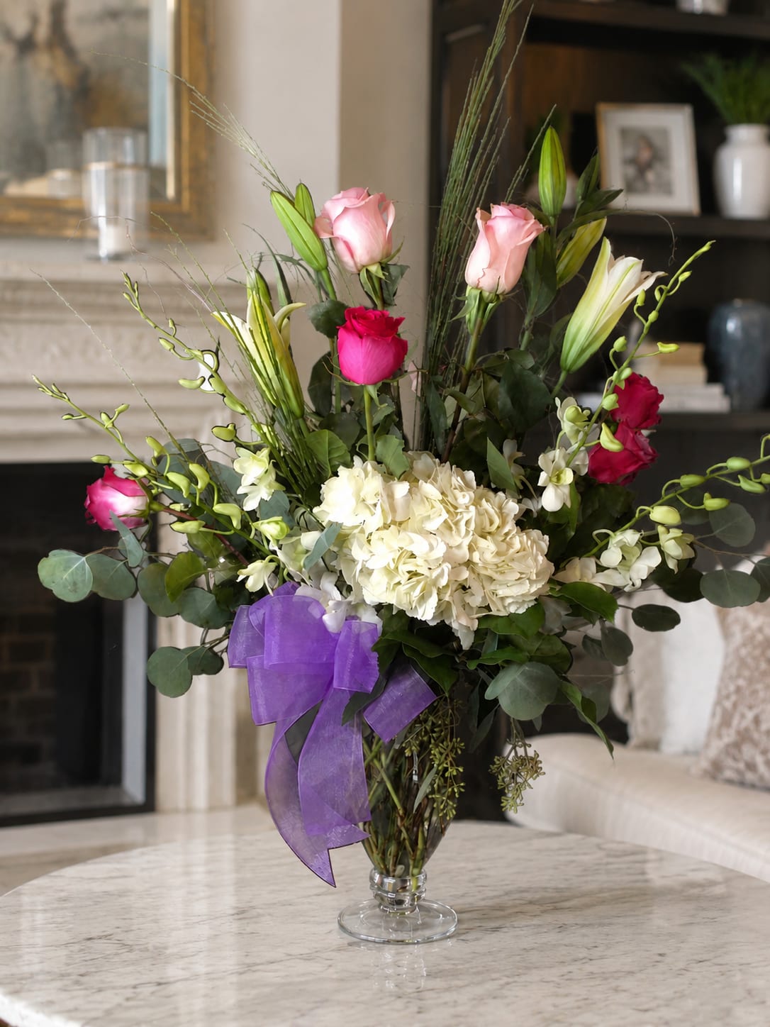 The Matriarch - A tall, layered flower arrangement that reads elegant and romantic, perfect as an anniversary bouquet, birthday surprise, or a "just because" gift to make her feel truly seen. This formal vase arrangement combines pale and bright pink roses with creamy white hydrangeas and white lilies, while arching orchid sprays create a graceful vertical silhouette-an ideal choice for weddings, graduations, or a thoughtful get-well present around Labor Day or any special moment.  This design is presented in a clear glass vase tied with a purple ribbon, with broad eucalyptus and airy foliage softening the structure for a polished, modern look. Expect long, layered stems and a mix of textures-lush hydrangea clusters, fragrant lilies, classic roses, and delicate orchids-giving the recipient a sense of luxury and care. Sized to command attention on an entry table or bedside, it's a meaningful way to say thank you, I'm sorry, congratulations, welcome new baby, or simply I love you; care instructions are included with your order to help keep blooms looking fresh.