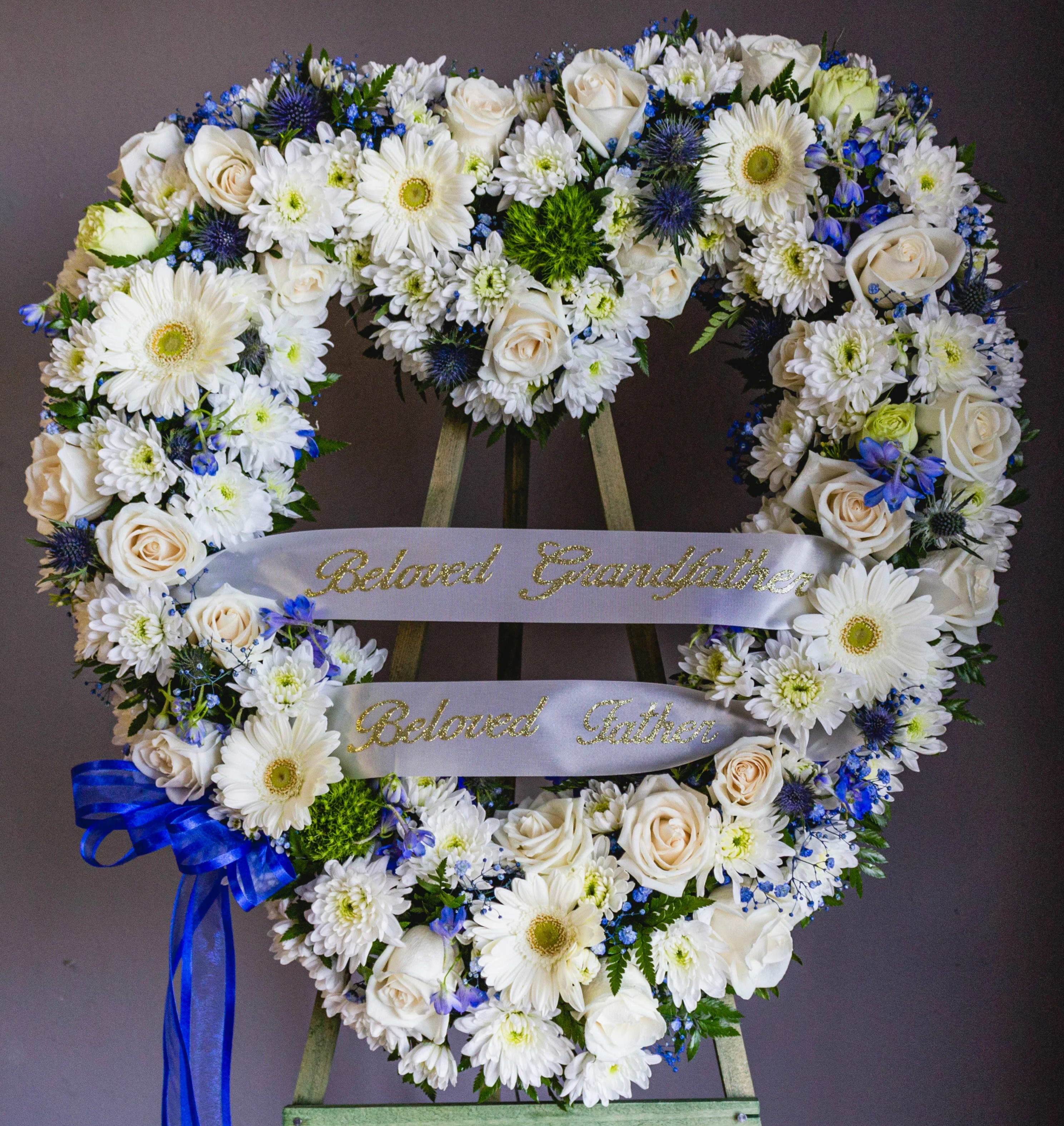 Heart-shaped floral wreath with white flowers and blue accents on a stand