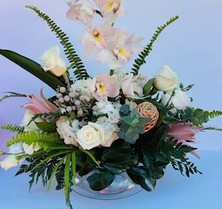 Bowl of exotics - Pale blush cymbidium orchid stems arch over a low clear glass bowl filled with soft white roses and creamy hydrangeas for a bowl-style flower arrangement that's perfect for an anniversary or a tender get-well wish. The design reads calm and refined-think understated luxury-so the person you're thinking of will feel genuinely comforted and celebrated whether this is sent for a birthday, a thank-you, or a sympathy moment.  Creamy white hydrangeas and roses create a lush, airy base while blush pink lilies and peachy accents add delicate height and movement. Prominent ferns and garden-like foliage give the arrangement a slightly asymmetrical, textural silhouette; the clear bowl vase keeps the focus on the blooms and works beautifully as a centerpiece for a bedside table or dining room. Sized to serve as an abundant, elegant table arrangement, it arrives ready to display-simply add fresh water and trim stems as needed to extend vase life.