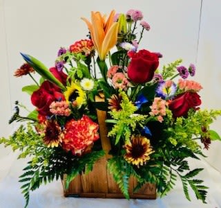 Rustic basket - Bright, cheerful color bursts of red, orange and sunny yellow make this rustic basket arrangement a joyful pick-me-up for birthdays, congratulations, or "just because" moments. The flower arrangement is a lively centerpiece-style floral gift that reads as celebratory and warm-perfect for the person you want to make feel noticed and genuinely cheered.  A full, upright mix of red roses, orange Asiatic lilies, multicolored carnations, chrysanthemums and daisy-style blooms is gathered in a rectangular wooden basket with a handle, creating a casual, country-chic presentation. Taller stems rise through the center while smaller blossoms cluster around the edges for a lush, abundant look; the varied textures-from smooth rose petals to ruffled carnations and open daisies-give the bouquet vibrant movement and visual interest.  This arrangement suits a sunny kitchen table, a cheerful office desk, or a bedside pick-me-up and sends a clear message of celebration or comfort. No special care is needed beyond fresh water-place in a cool spot away from direct sun and change the water every other day to keep blooms looking their best.