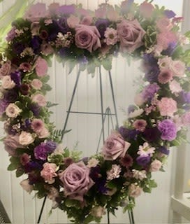 Open Heart wreath - pick your palette - A heart-shaped sympathy wreath composed in gentle lavenders and pinks-designed to offer a tender, reflective tribute at a memorial or funeral. The Lavender heart wreath reads as a thoughtful expression of love and remembrance, the kind of floral tribute that lets the person you're honoring feel truly seen and cherished in a solemn moment.  Densely covered in lavender roses with soft pink accents and richer purple blooms, the wreath forms a continuous, full ring of texture and color. Carnations and chrysanthemums are interwoven with mixed bouquet blooms to create soft transitions from pale pastels to deeper tones, giving the piece a layered, comforting presence. The arrangement is mounted on a wreath frame and shown on a simple metal easel stand for upright display, making it suitable for service chapels, graveside presentations, or a memorial corner.  For someone arranging a service or sending condolences, this wreath conveys sympathy with quiet elegance. It arrives ready for display on the included easel; no additional assembly is required. The muted palette and classic heart shape make it an appropriate, lasting way to express love and remembrance.