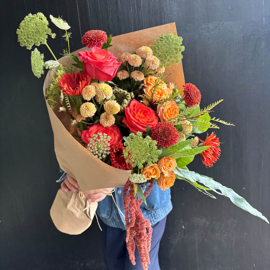 Handheld bouquet of pink, orange, and red flowers wrapped in brown paper