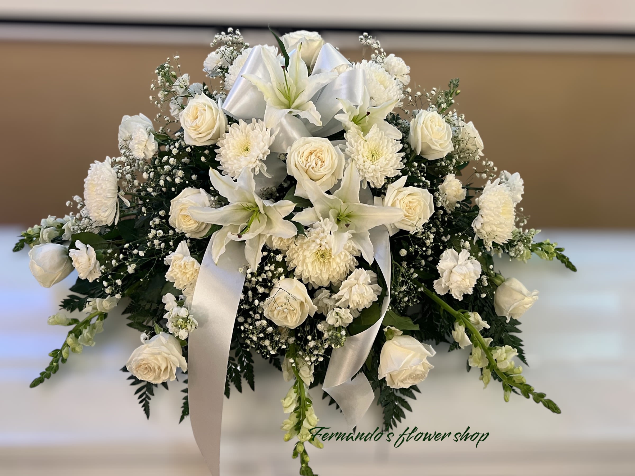 Low white arrangement of lilies, roses, carnations, and mums with a white ribbon bow