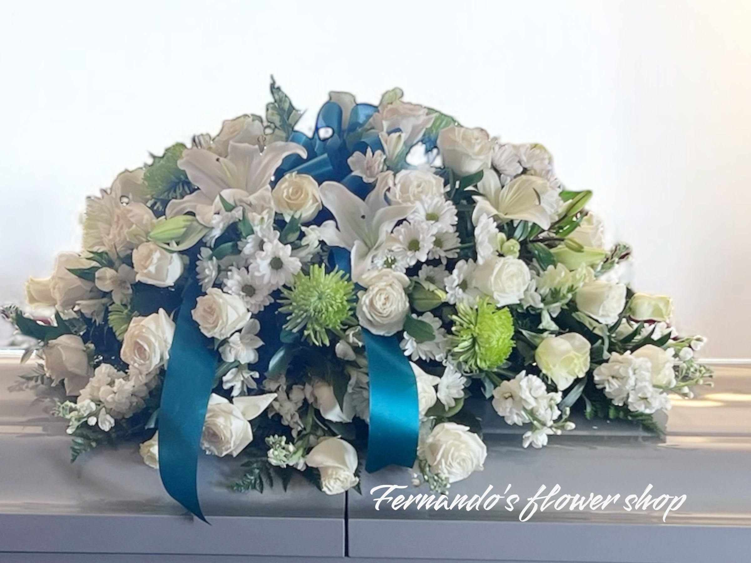 White and green casket spray of roses, lilies, and mums with teal ribbon on a white casket.