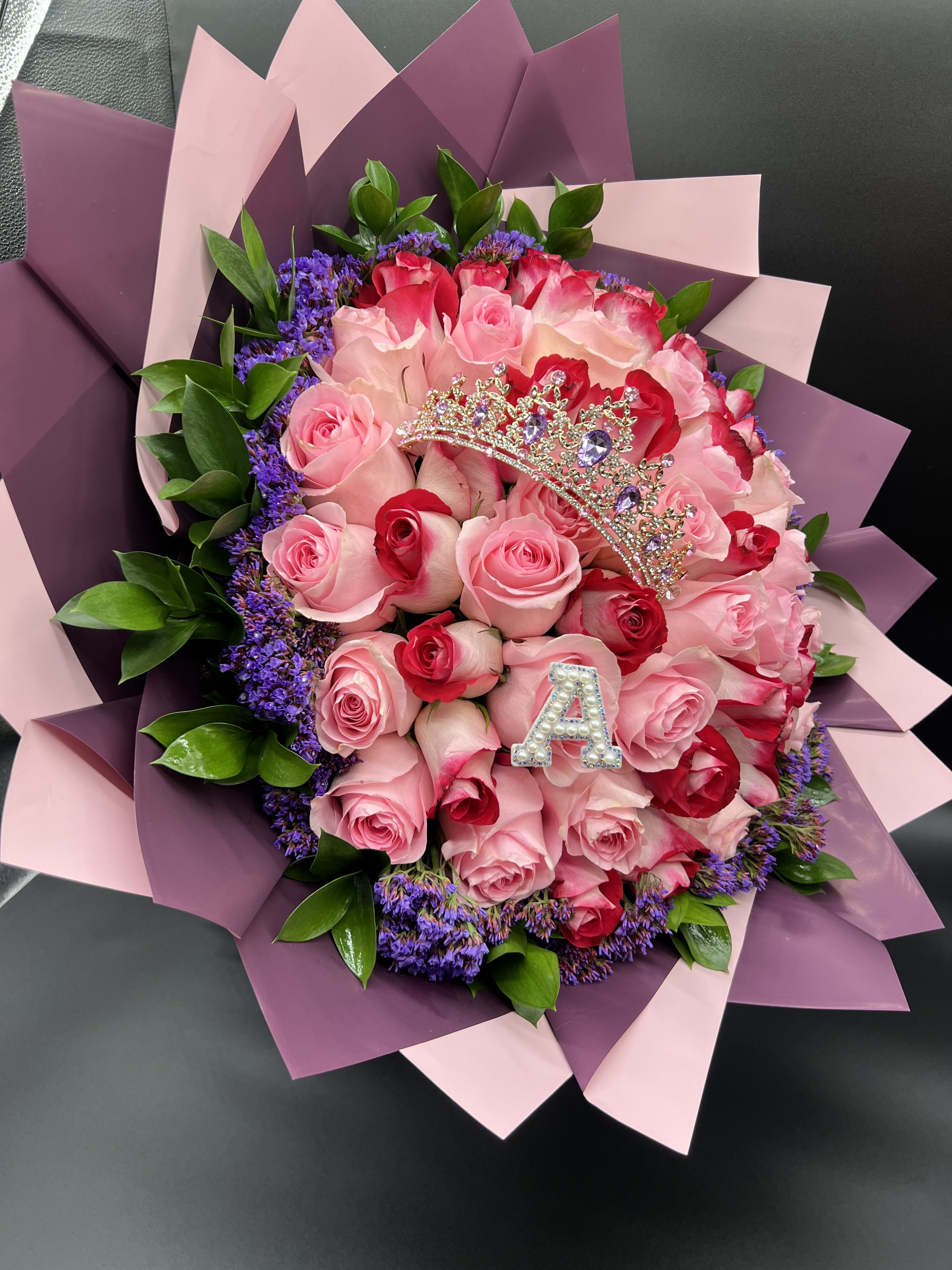 Bouquet of pink and red roses with purple filler flowers, wrapped in pink paper with a jeweled tiara and letter A.