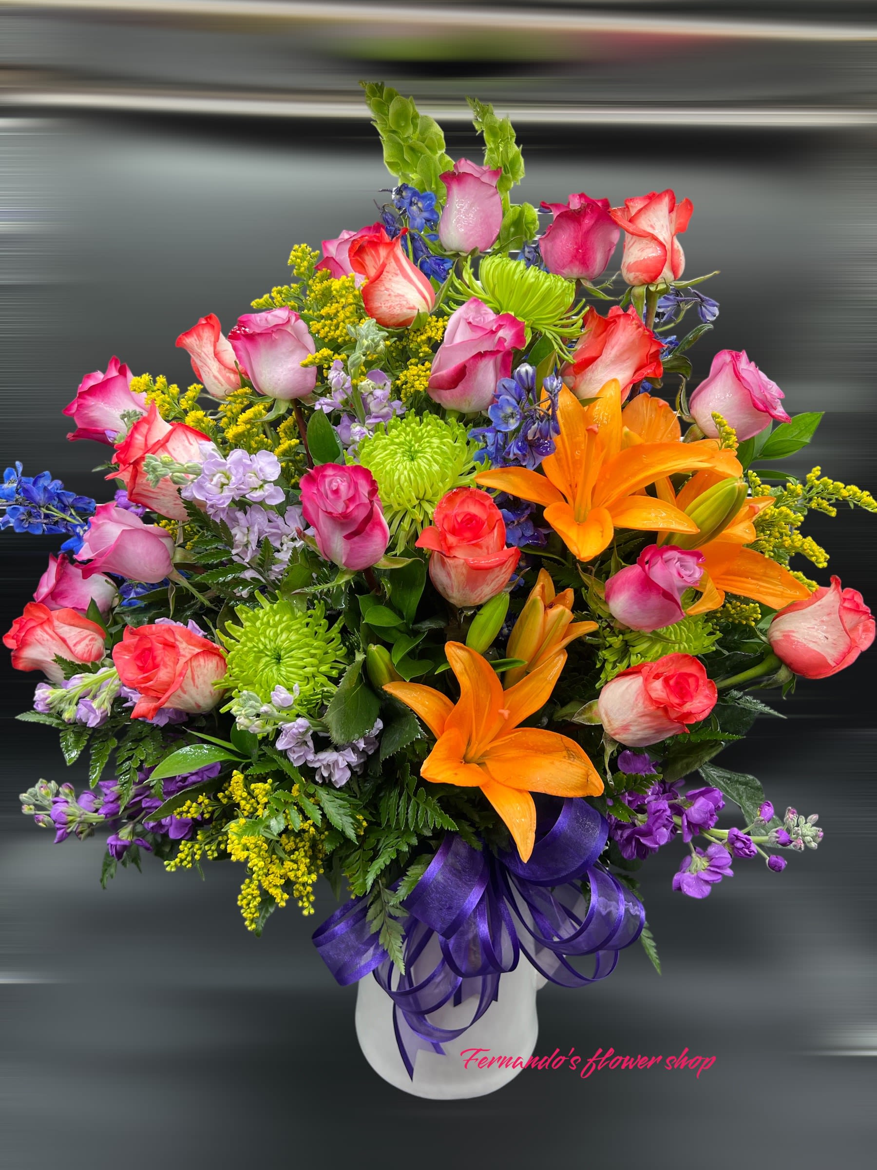 Tall mixed arrangement of pink roses, orange lilies, green mums, and blue accents in a white vase with purple ribbon.