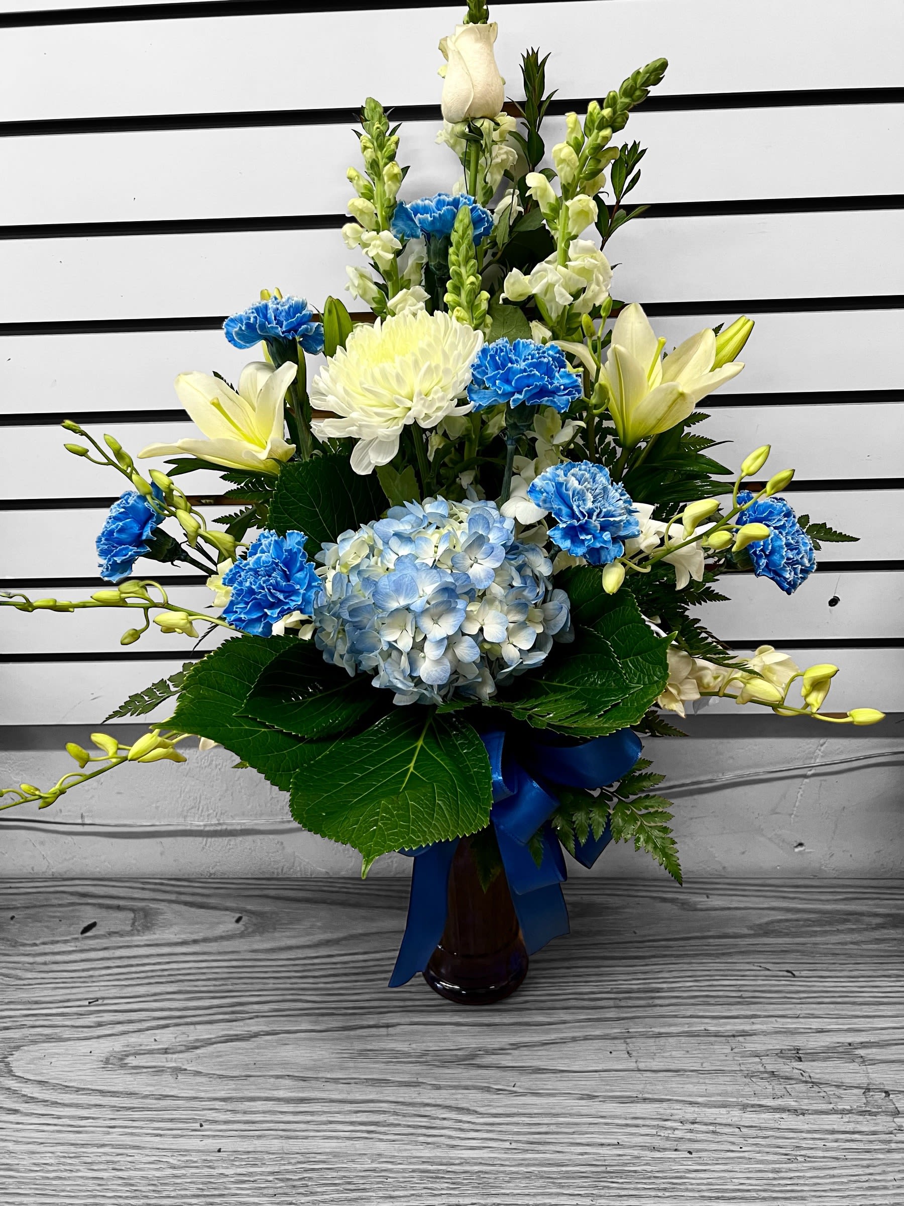 Tall blue and white flower arrangement with lilies, carnations, and hydrangea in a vase with a blue ribbon