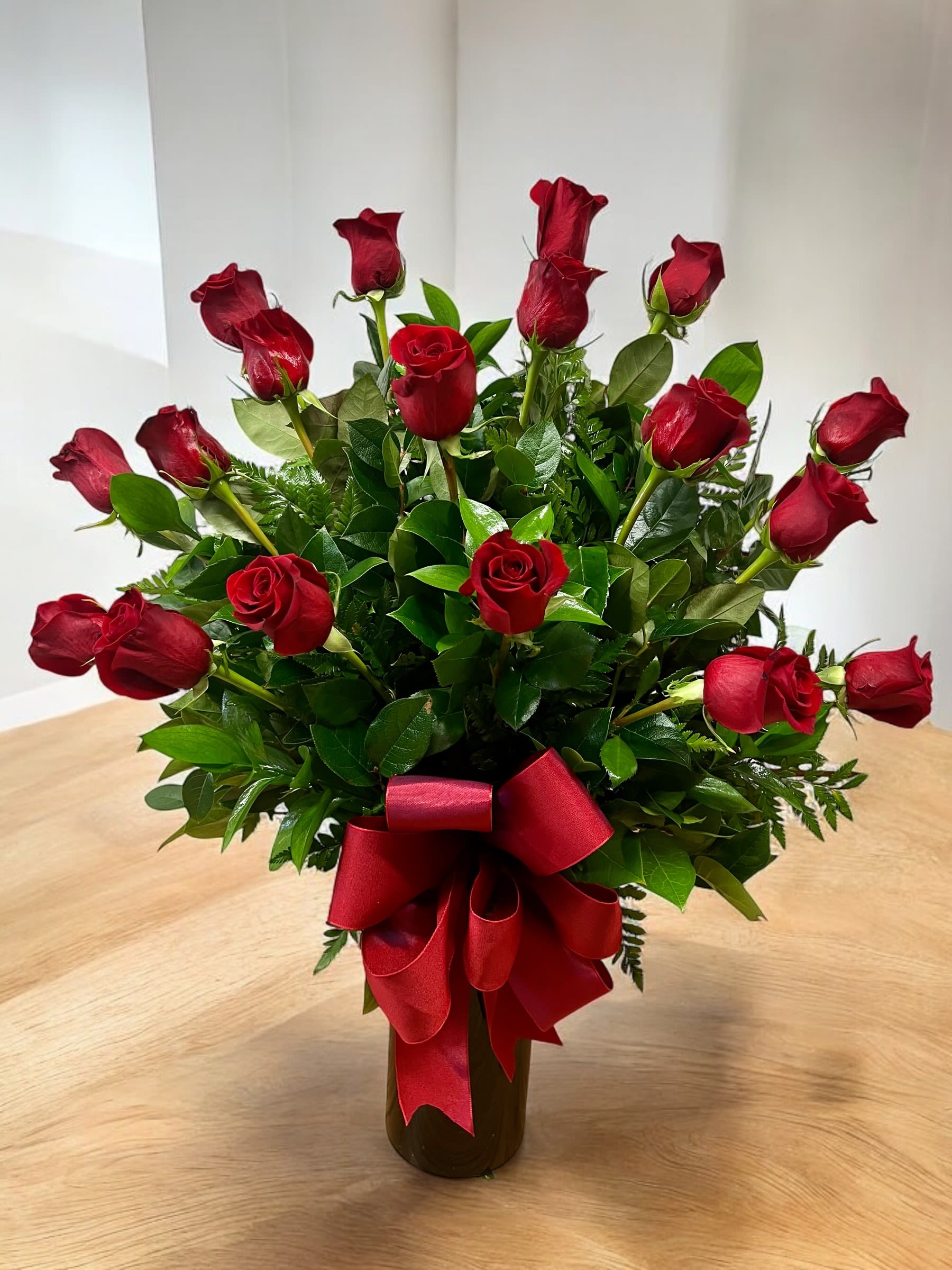 Tall arrangement of long-stem red roses with lush foliage in a dark vase with a red bow