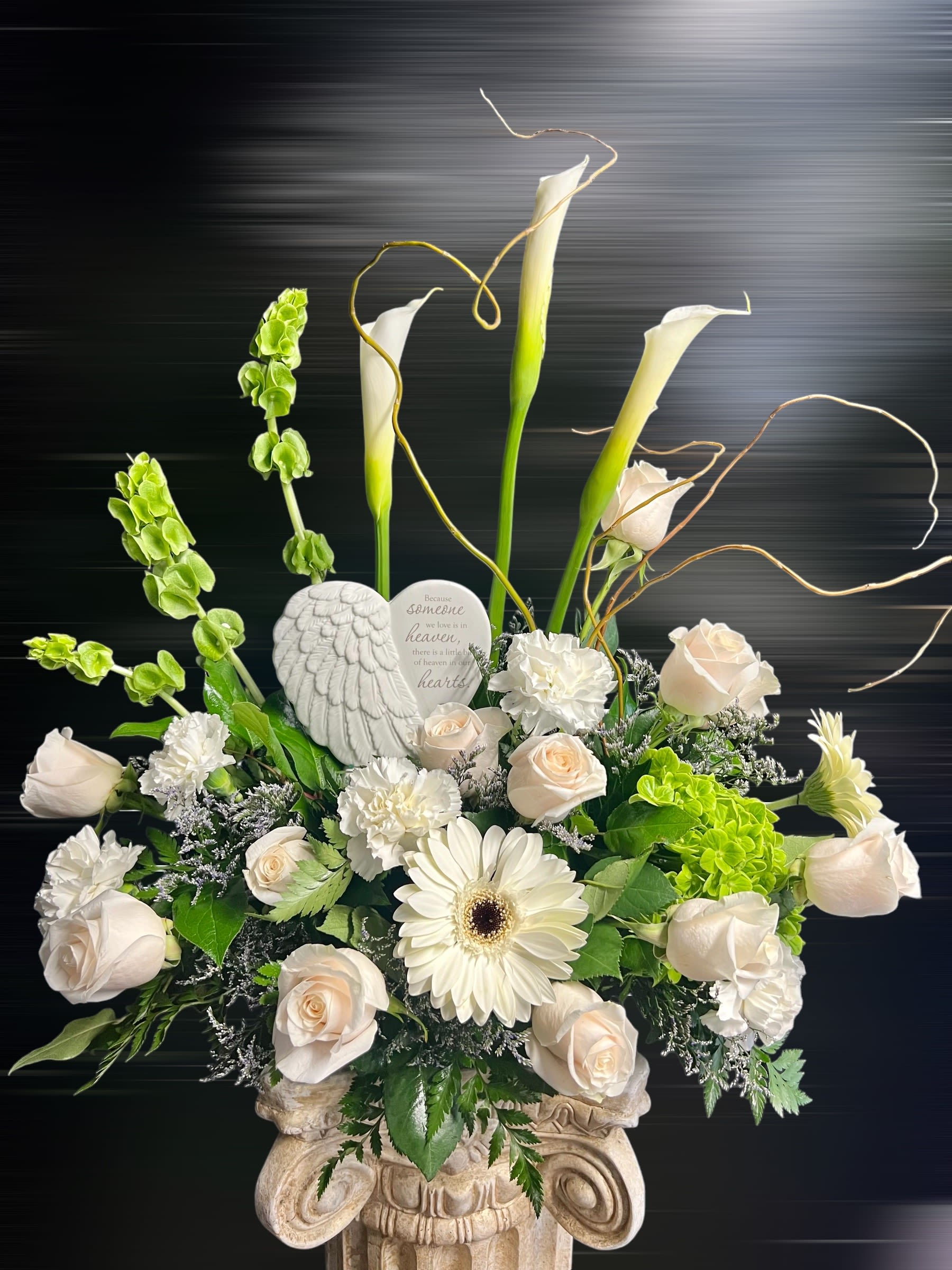Tall white and green sympathy arrangement with roses, calla lilies, and angel plaque in a stone urn.