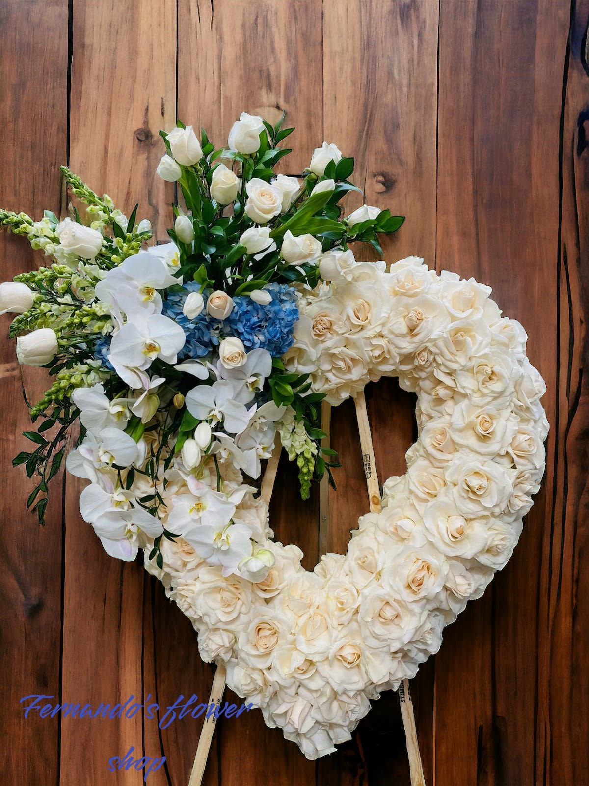 Heart-shaped ivory rose wreath with white orchids and blue hydrangeas on a wooden easel.