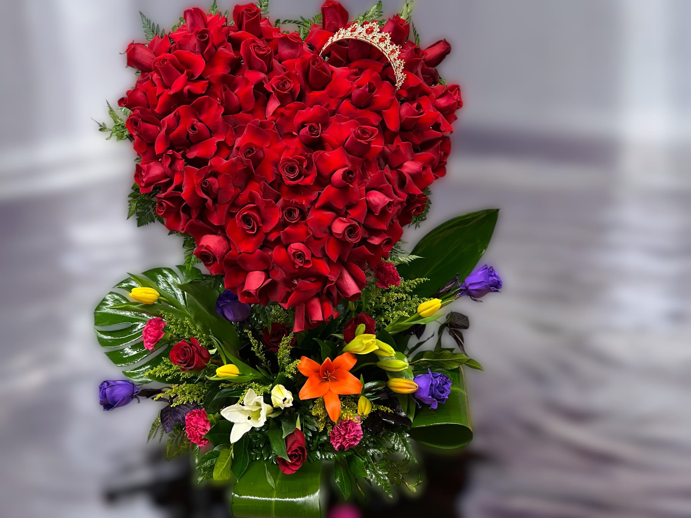 Tall arrangement with a heart of red roses above mixed colorful blooms and a gold tiara accent.
