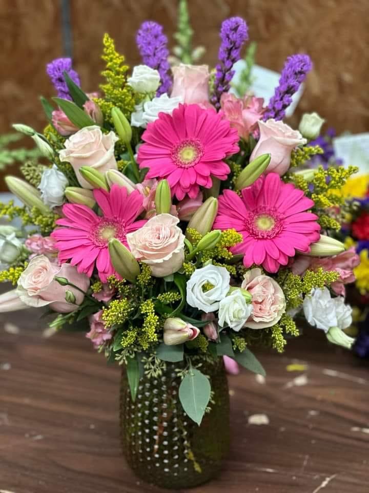 Vase arrangement with hot pink gerbera daisies, pale roses, lilies, and mixed blooms in a green textured glass vase