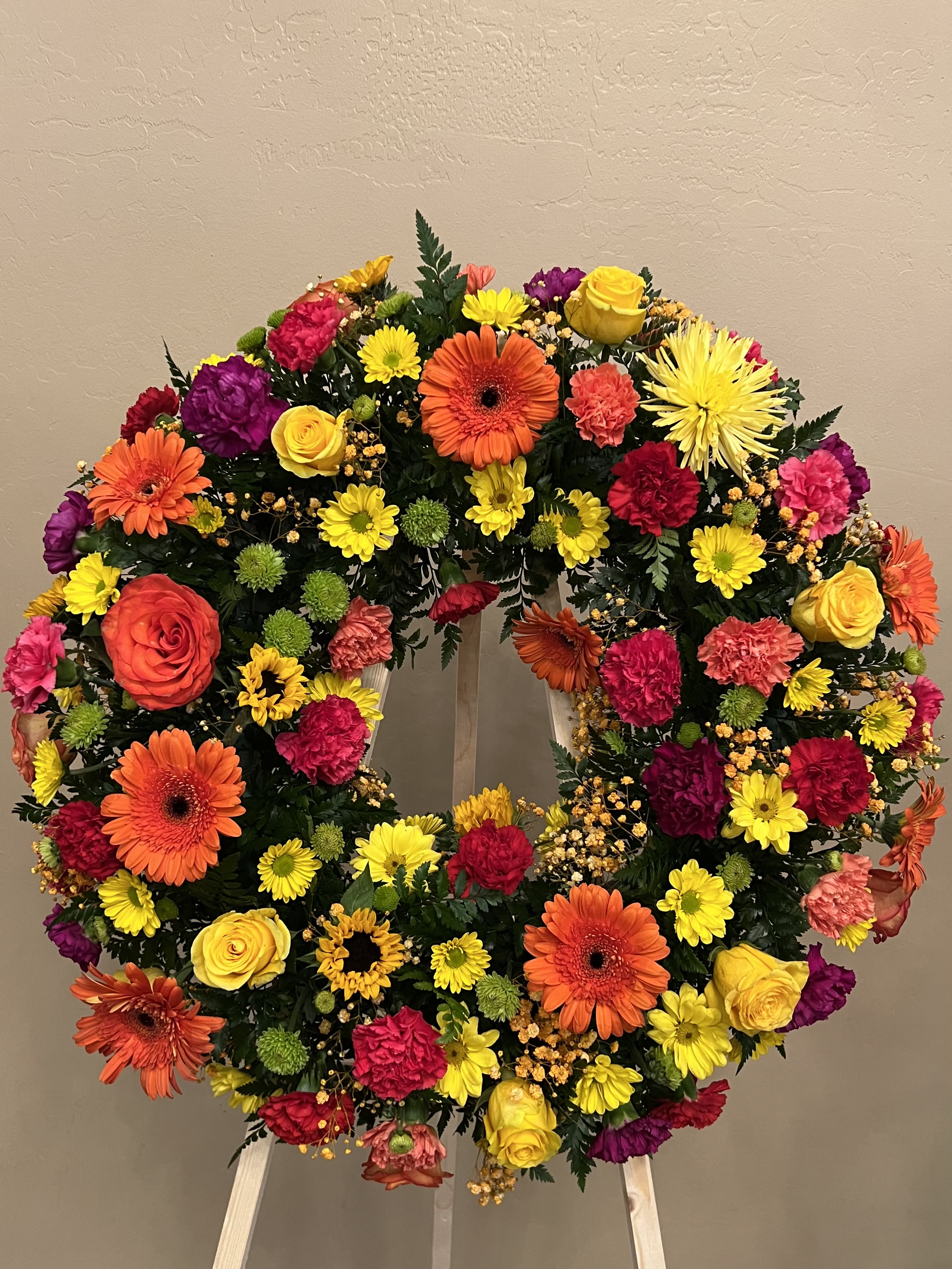 Bright circular wreath of orange gerbera daisies, yellow roses, and mixed flowers on a wooden easel.