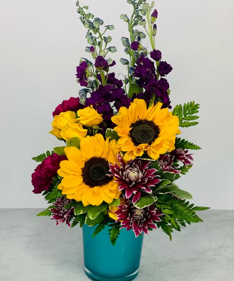 Tall arrangement with sunflowers, purple blooms, and bicolor mums in a turquoise vase