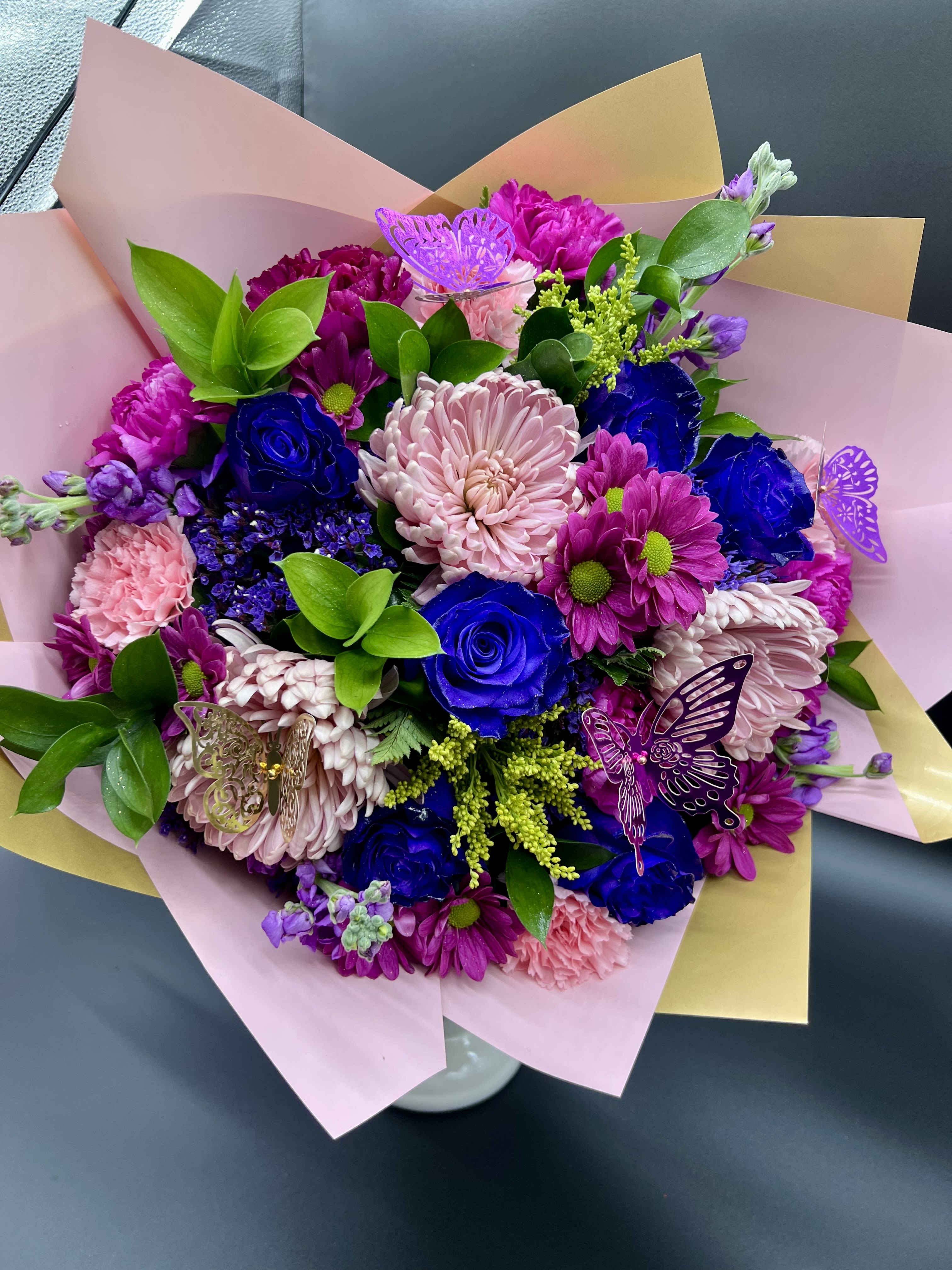 Hand-tied bouquet of pink mums and blue roses wrapped in pink and yellow paper with butterfly picks