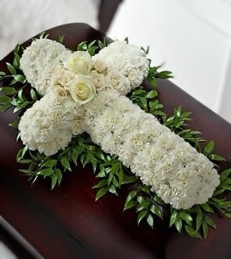 White floral cross with roses and carnations on greenery displayed on a dark wood casket