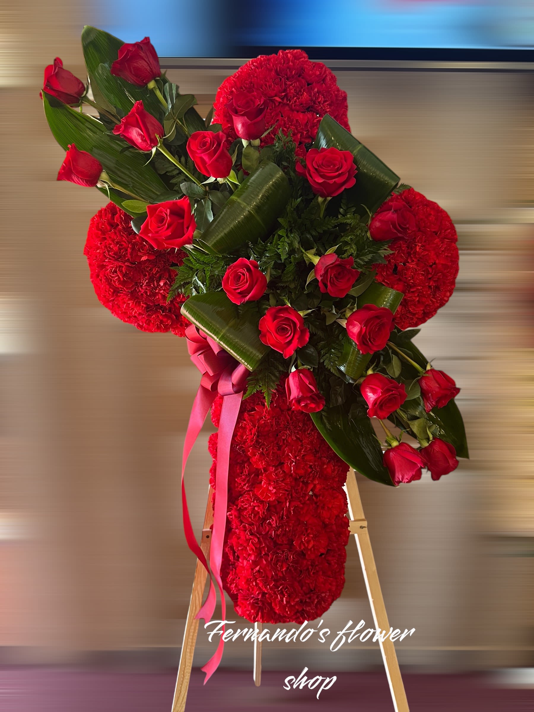 Standing red floral cross with red roses, large green leaves, and ribbon on a wooden easel.