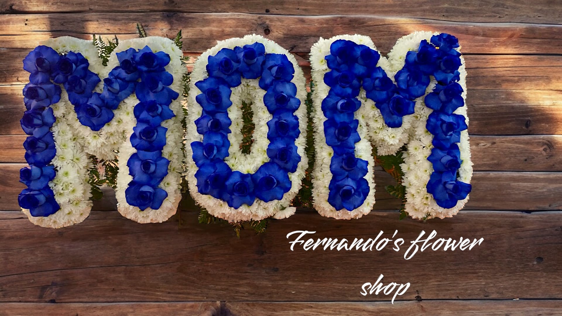 Floral letters spelling MOM with blue roses on white flowers against wood background