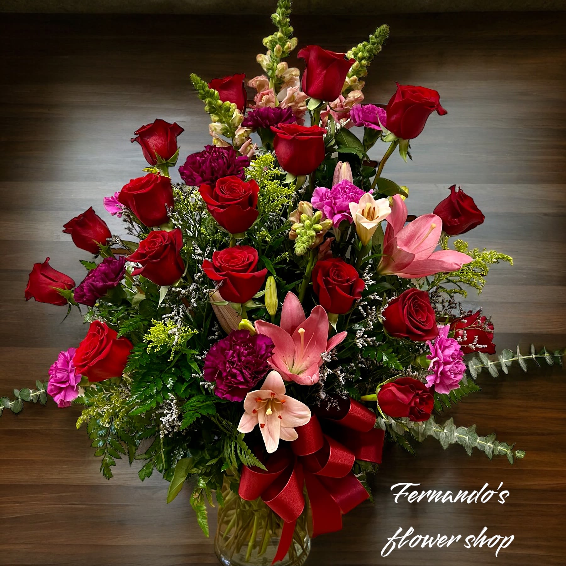Tall arrangement of red roses, pink lilies, and carnations in a clear vase with a red bow