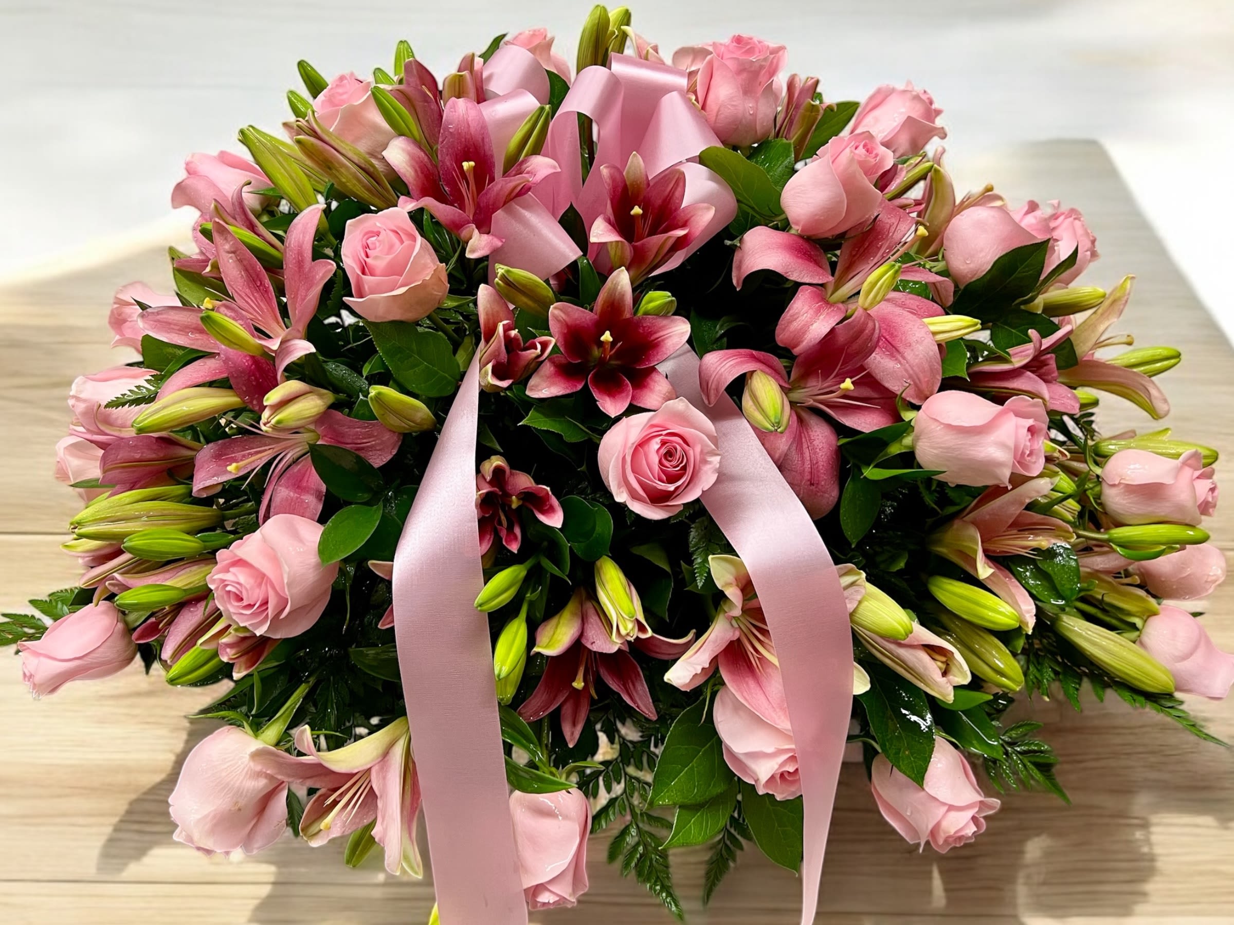 Low arrangement of pink roses and pink lilies with buds, accented by a large pink ribbon bow.