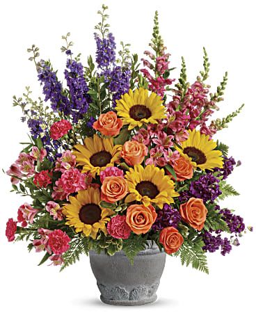 Twilight Bloom - This vibrant arrangement bursts with the beauty of a sunny garden. Featuring bright yellow sunflowers, cheerful orange roses, delicate pink carnations, and bold purple blooms like larkspur and snapdragons, this display is a celebration of color and life. Accented with lush greenery and arranged in a charming ceramic vase with subtle floral detailing, it exudes warmth, happiness, and positivity. Perfect for brightening someone’s day, celebrating special occasions, or simply bringing joy to any space.