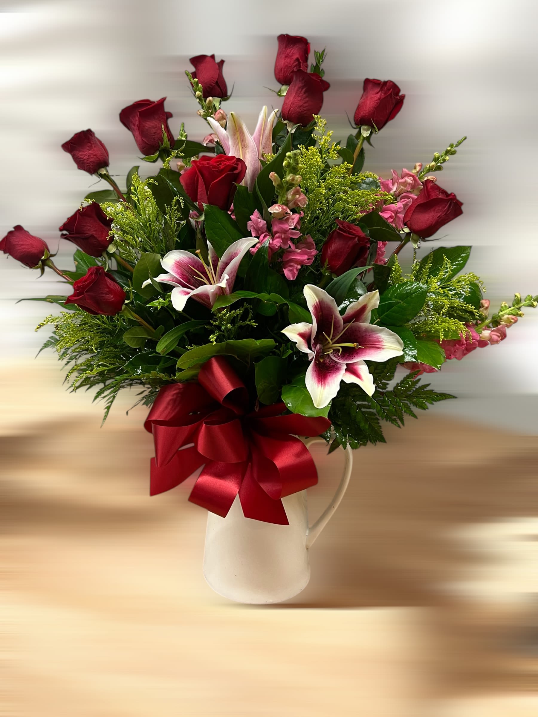 Tall arrangement of red roses and pink lilies in a white pitcher with a large red bow