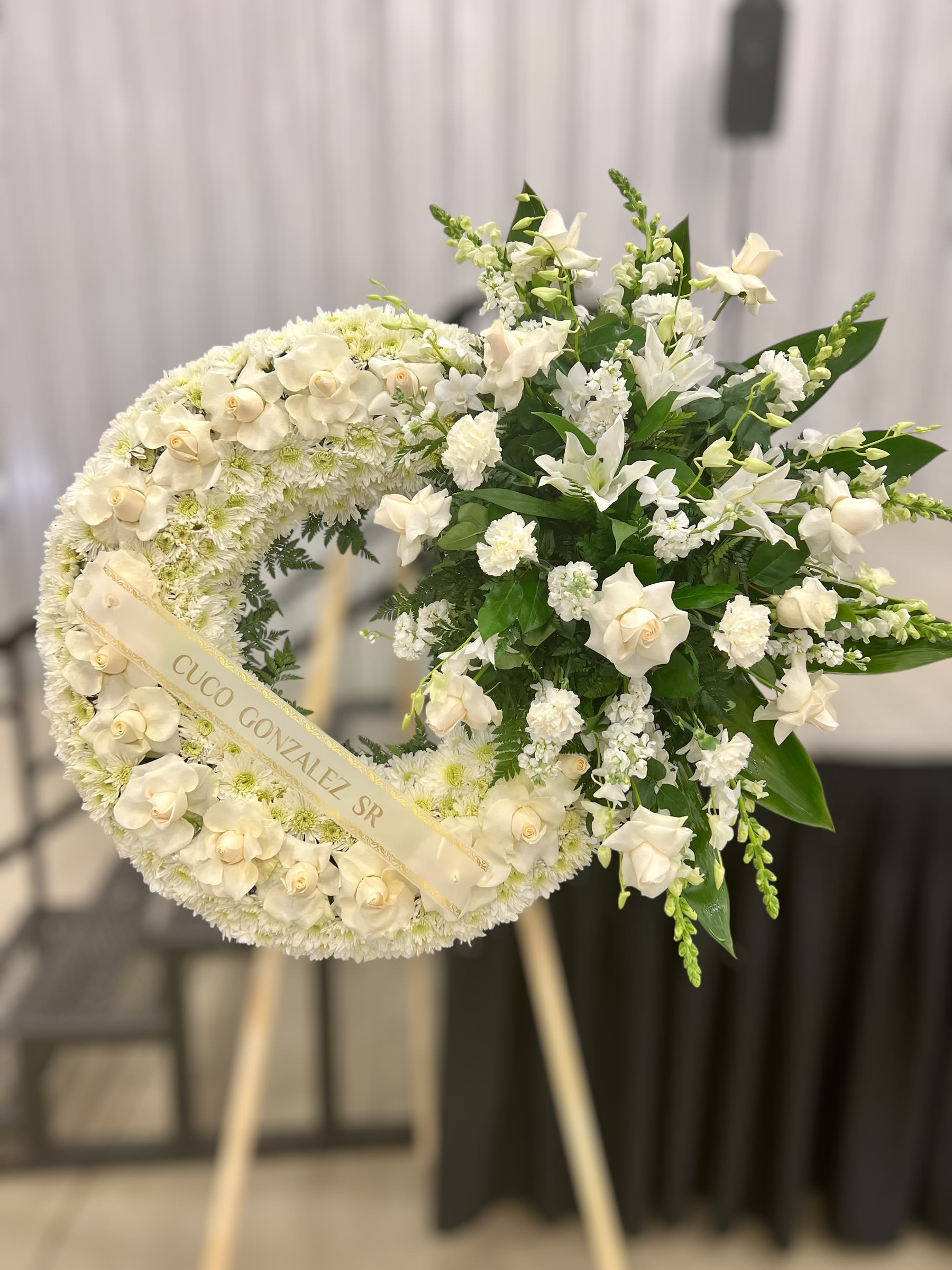 Standing white funeral wreath with roses and lilies on an easel with name ribbon