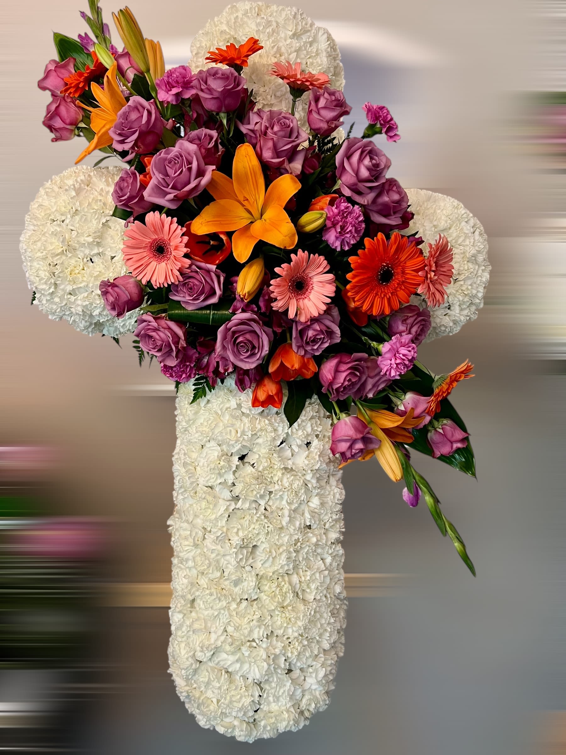 Standing white floral cross with cluster of purple roses, orange lilies, and pink gerberas.