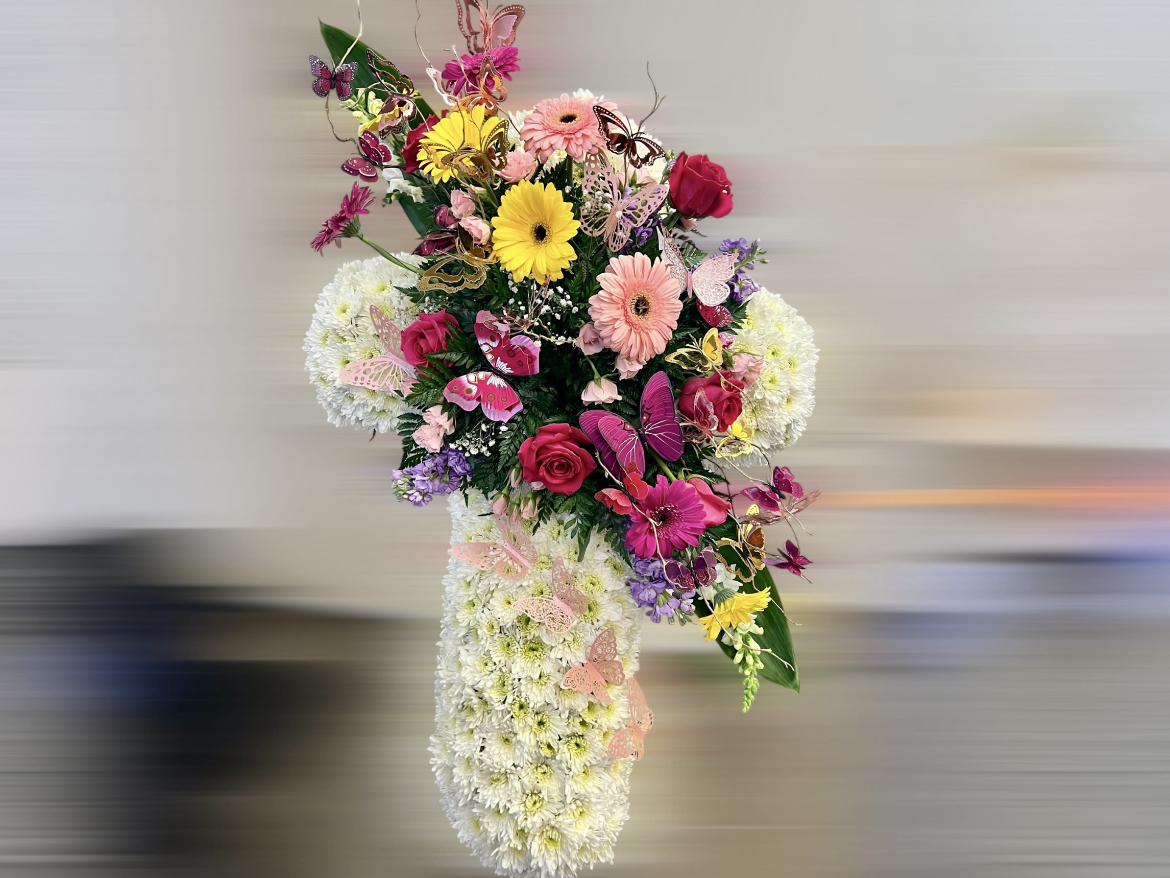 Floral cross of white mums adorned with pink roses, gerberas, lilies and colorful butterfly accents.