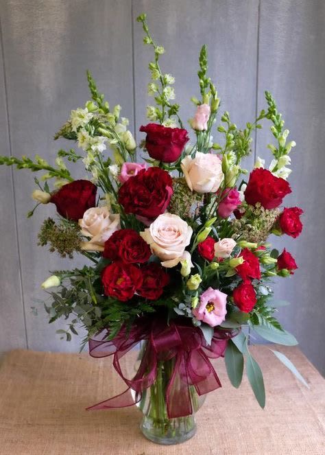 Tall arrangement of red and blush roses with pink blooms and white snapdragons in a glass vase with burgundy bow