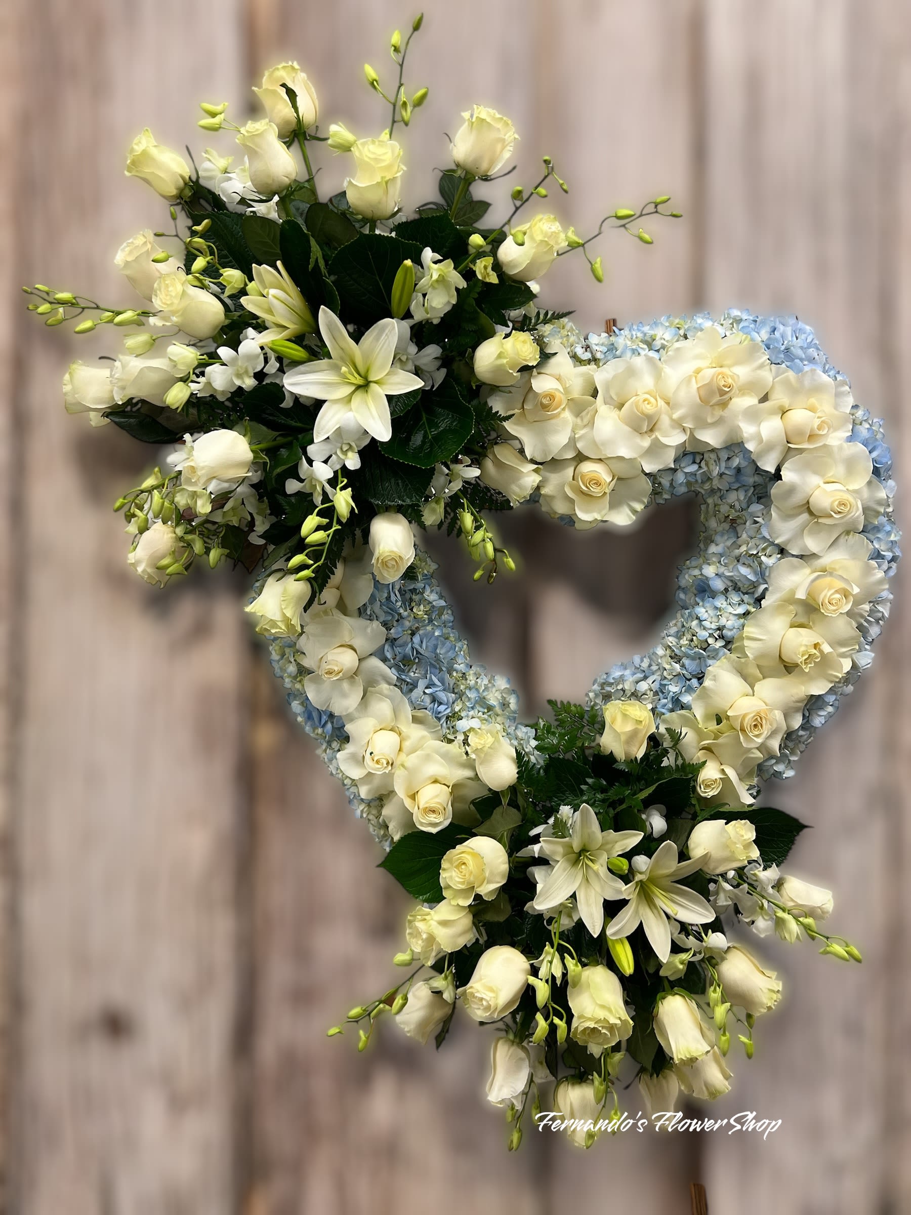 Heart-shaped wreath of white roses, lilies, and light blue hydrangeas on a stand
