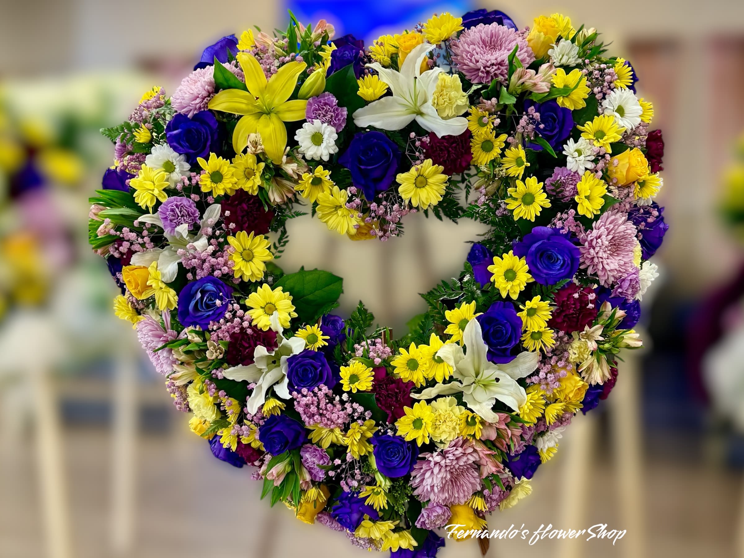 Heart-shaped sympathy wreath with yellow lilies, blue roses, daisies, and mixed flowers.