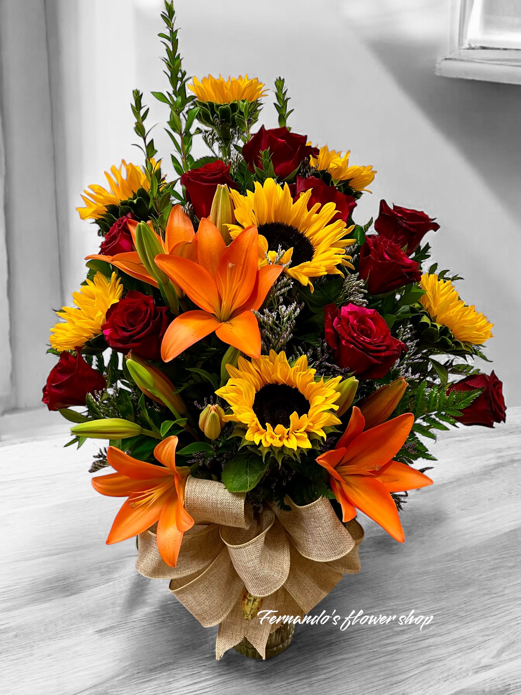 Mixed arrangement of sunflowers, orange lilies, and red roses in a glass vase with a burlap bow