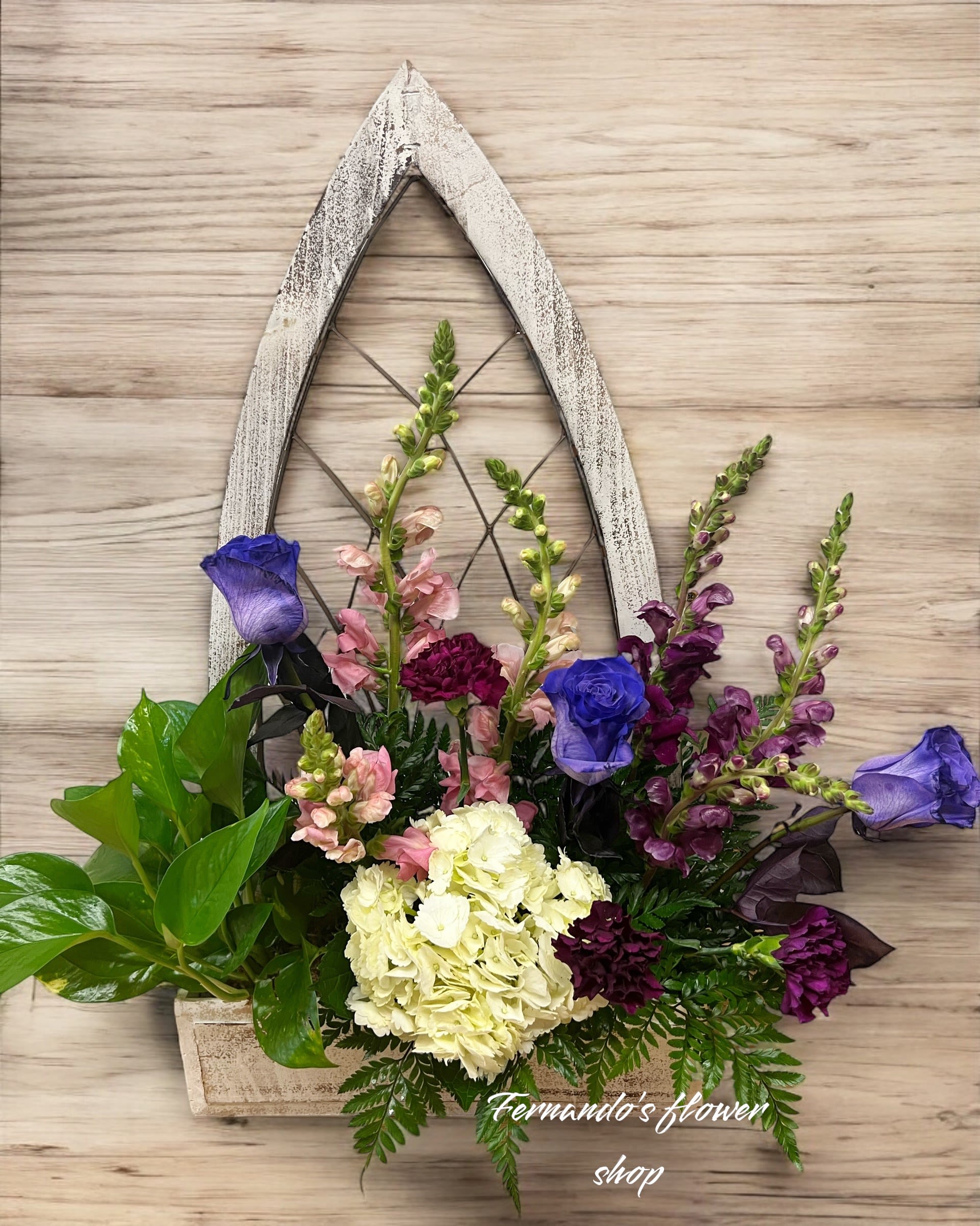 Wall-mounted rustic wooden frame planter with purple roses, white hydrangea, snapdragons, carnations, and green foliage.