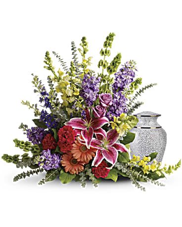Sympathy floral spray of lilies, roses, and mixed flowers arranged beside a decorative cremation urn.