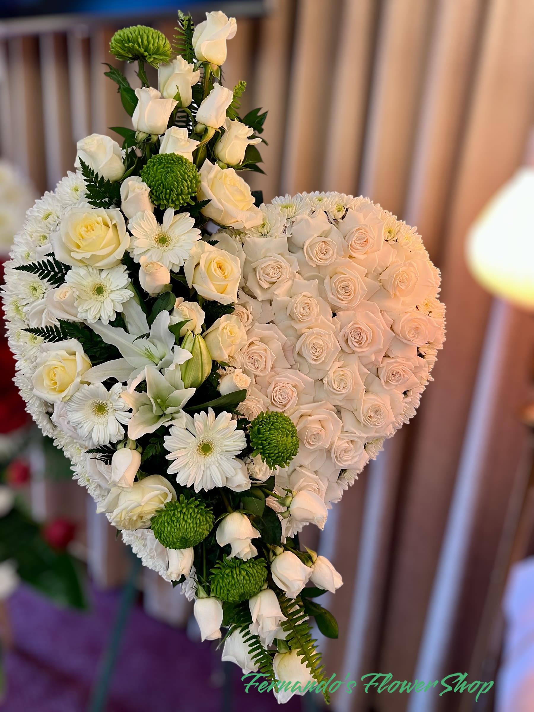 Standing heart-shaped spray of white roses, daisies, and lilies with green accents on an easel.