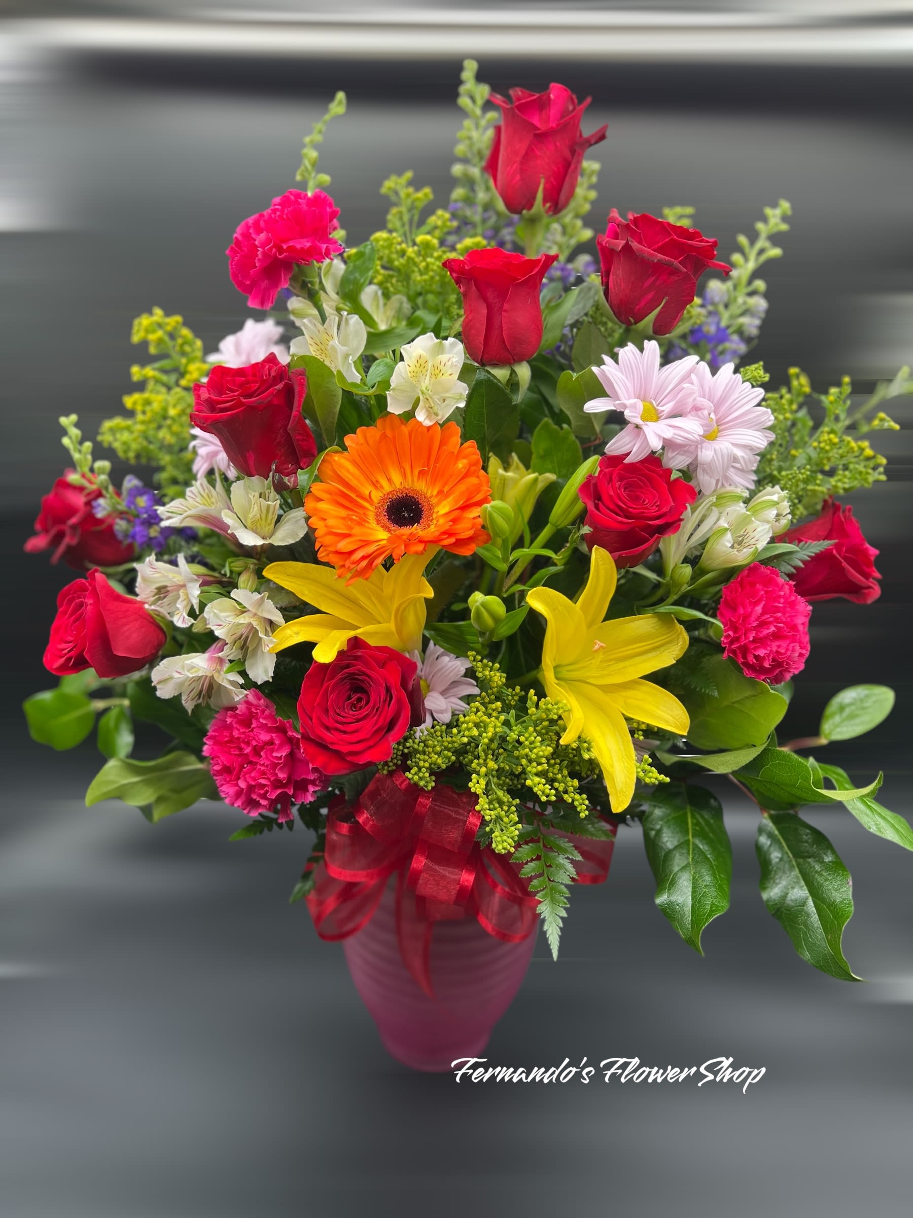 Mixed arrangement of red roses, yellow lilies, pink carnations and orange gerbera in a pink vase