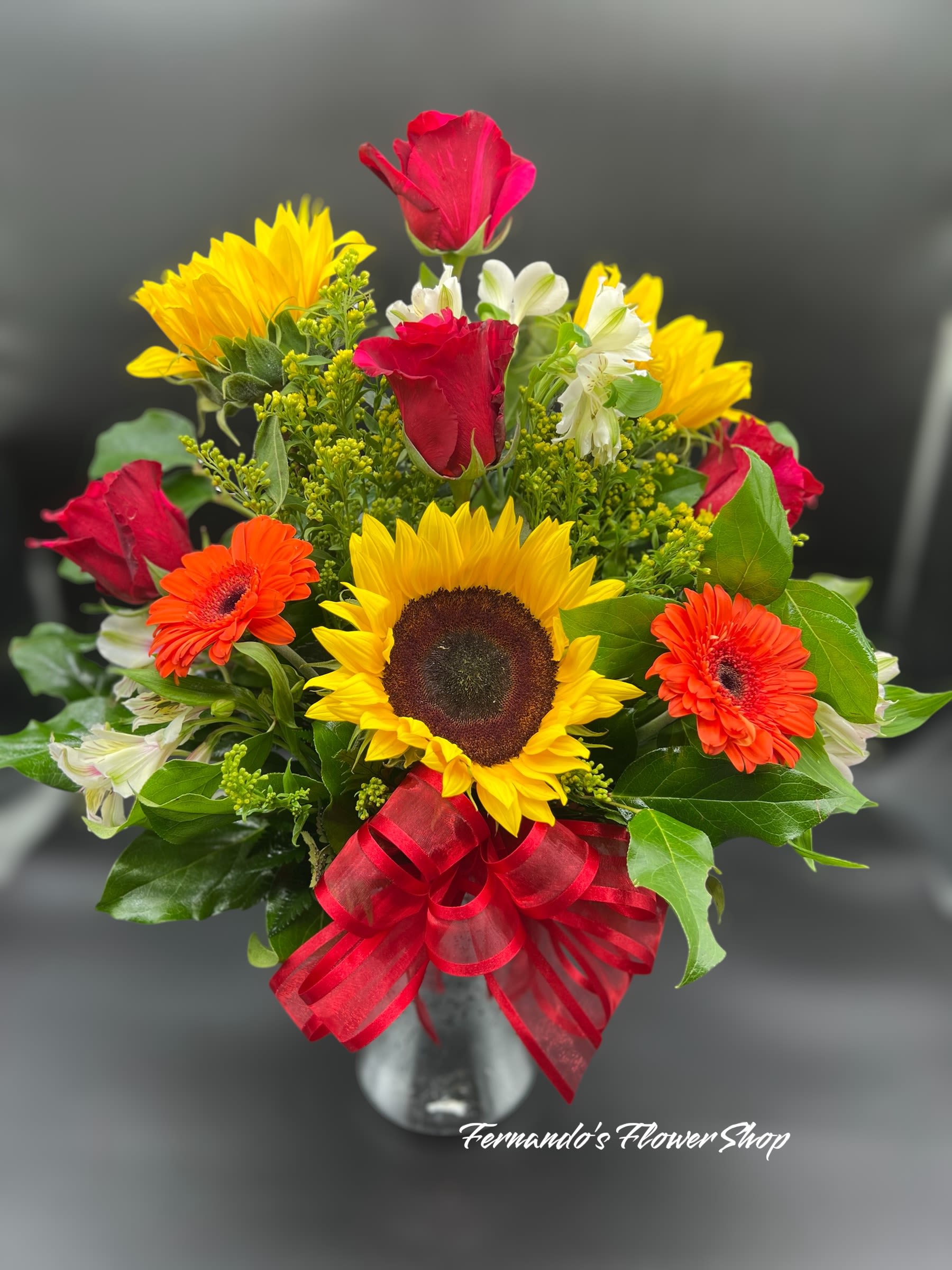 Bright mixed arrangement with sunflowers, red roses, orange gerberas and white blooms in a glass vase with red bow