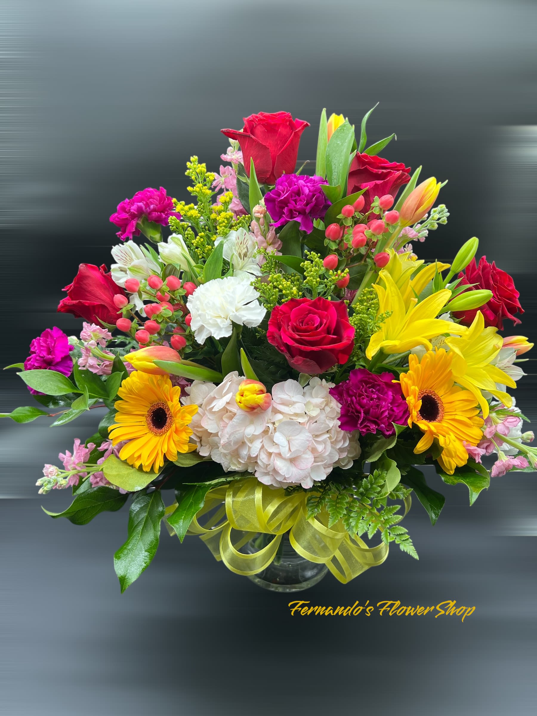Bright mixed flower arrangement with red roses, yellow lilies, and gerbera daisies in a glass vase.