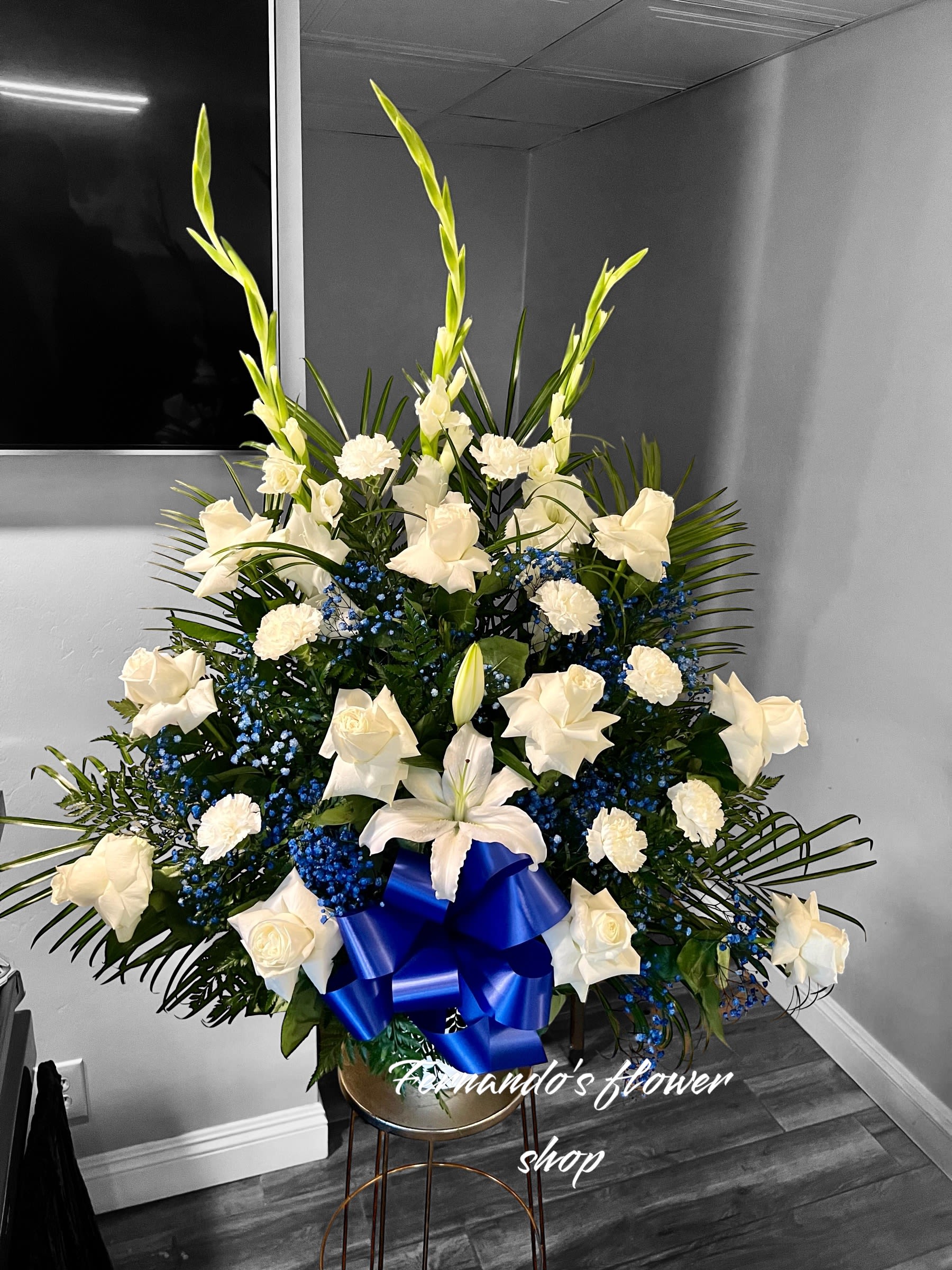 Tall white and blue floral arrangement with roses, lilies, and carnations in a container with a blue bow