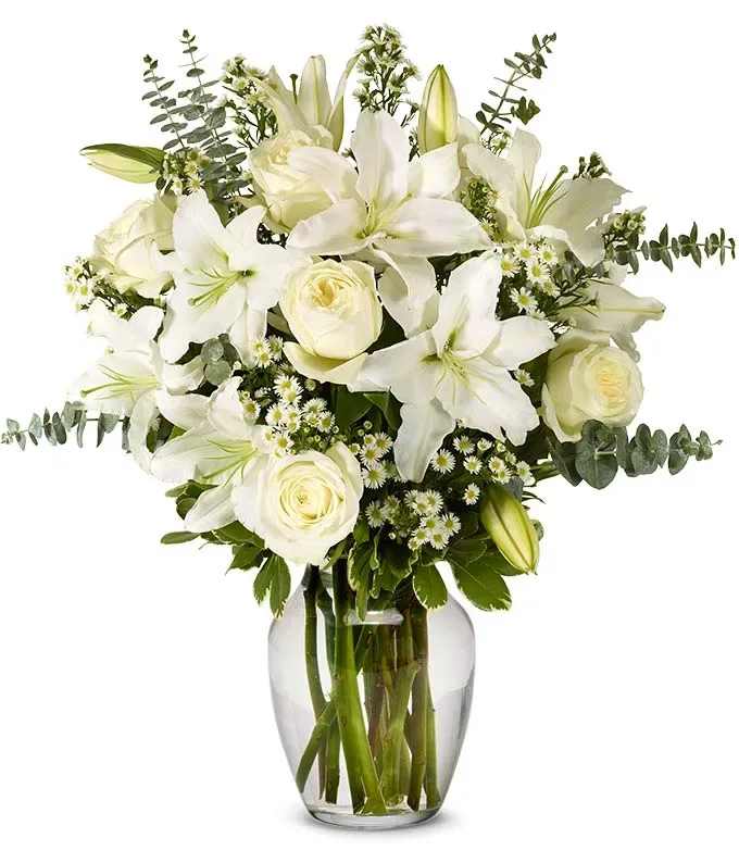 White lilies, roses, and small white blossoms arranged in a clear glass vase