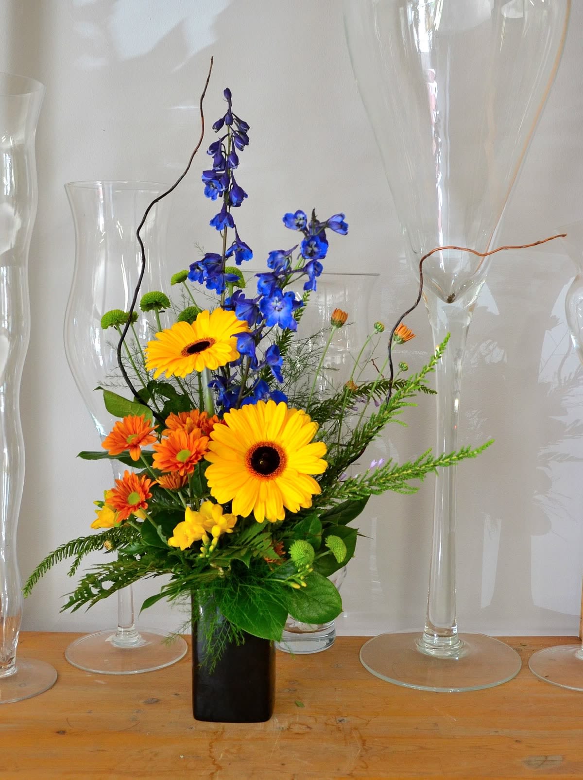 Tall arrangement of yellow gerbera, orange mums, and blue delphinium in a black rectangular vase