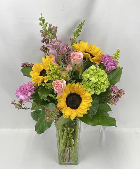 Tall mixed bouquet with sunflowers, pink roses, purple blooms, and green hydrangea in a clear glass vase