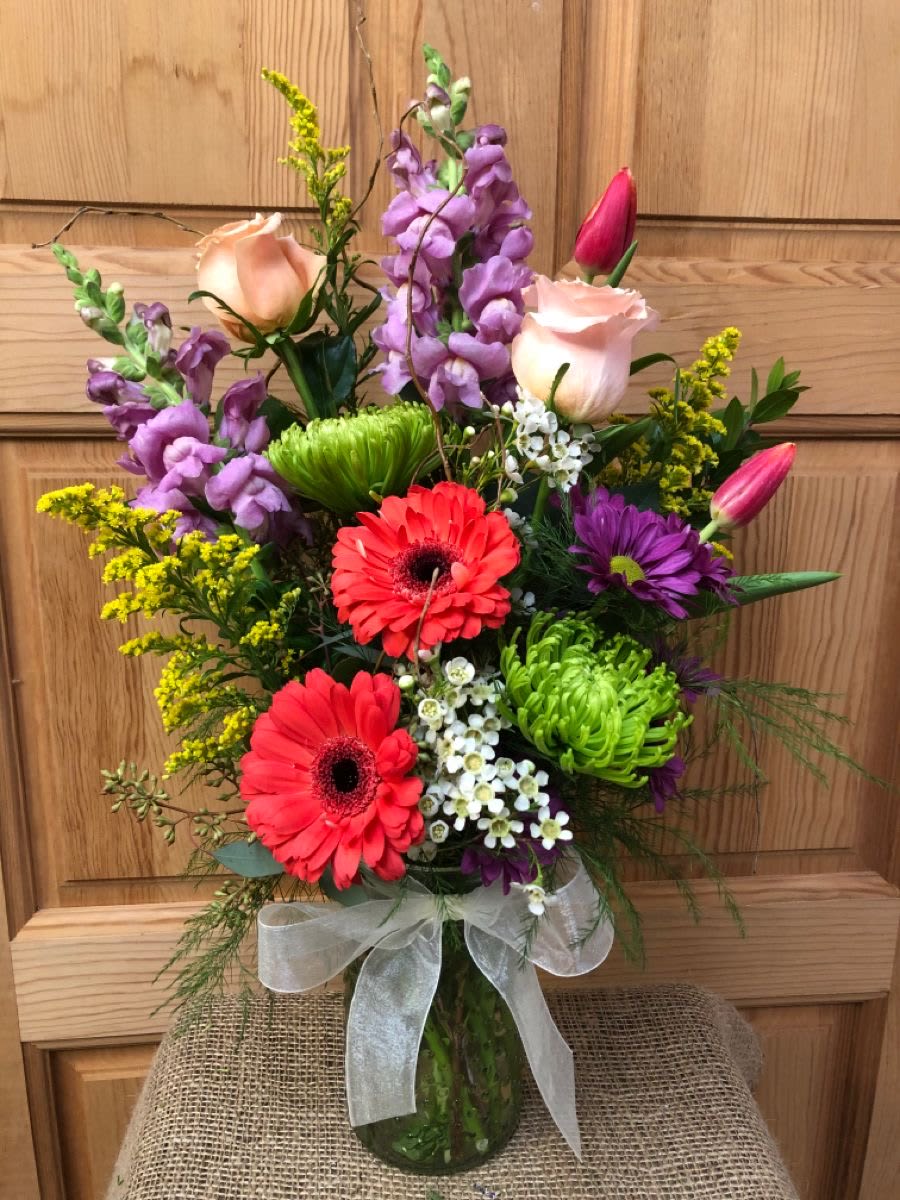 Mixed bouquet with pink roses, red gerbera daisies, green mums, and tulips in a glass vase with ribbon