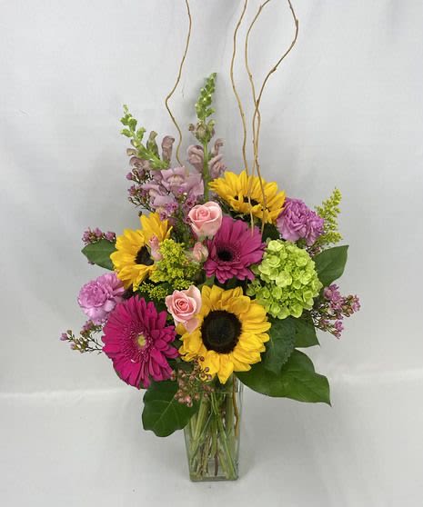 Tall mixed arrangement with sunflowers, pink gerberas, roses, and green hydrangeas in a clear glass vase