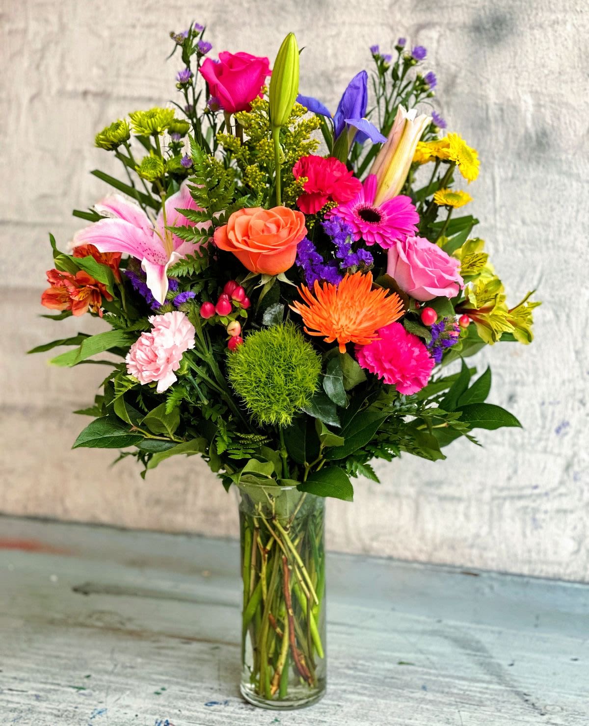 Tall mixed bouquet of bright pink, orange, yellow, and purple flowers in a clear glass vase.
