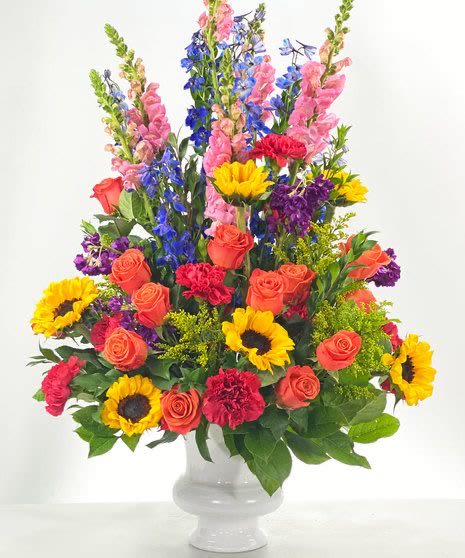 Tall mixed arrangement of sunflowers, orange roses, pink snapdragons and purple flowers in a white vase