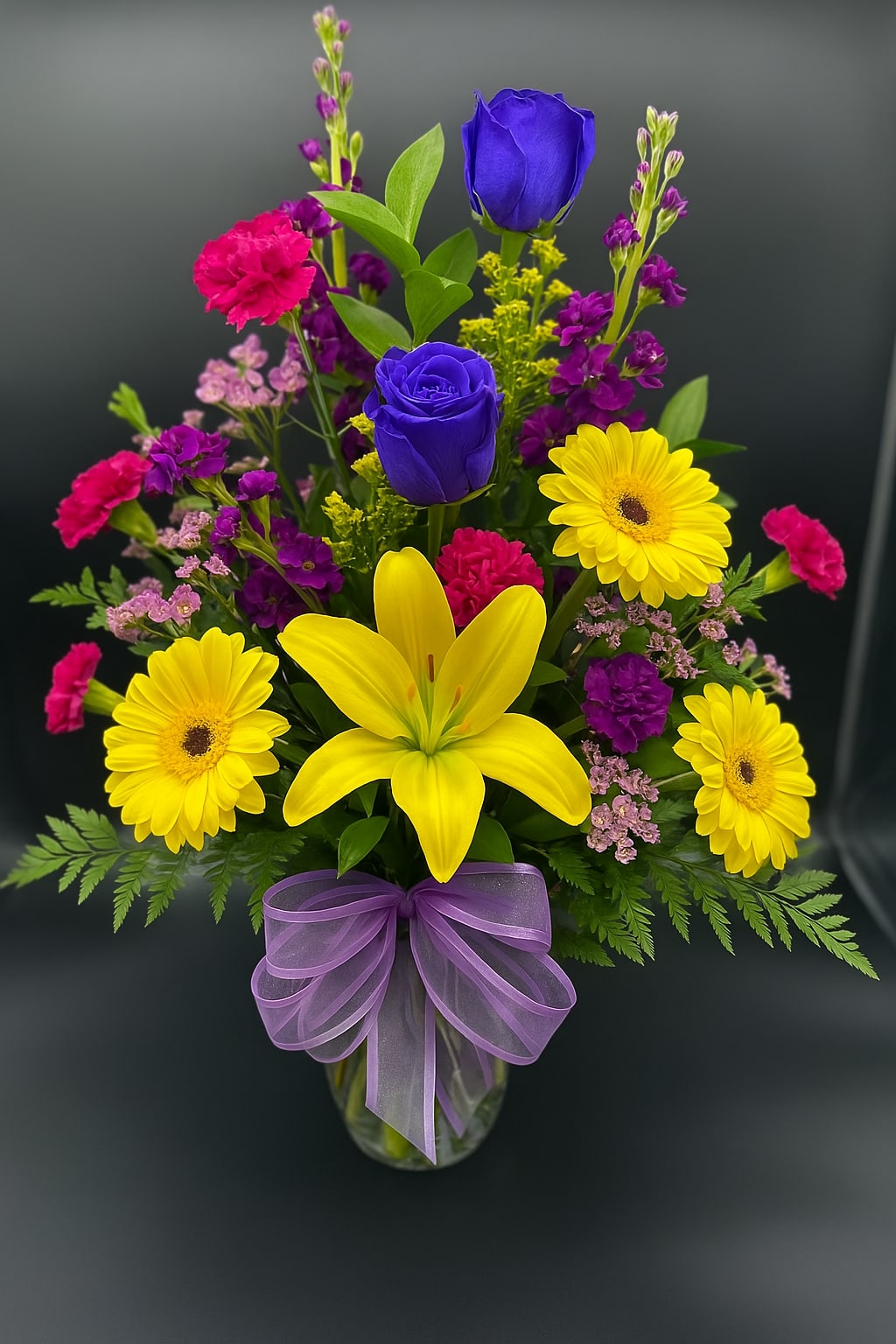Tall mixed bouquet with yellow daisies and lily, blue roses, pink carnations in glass vase with purple bow