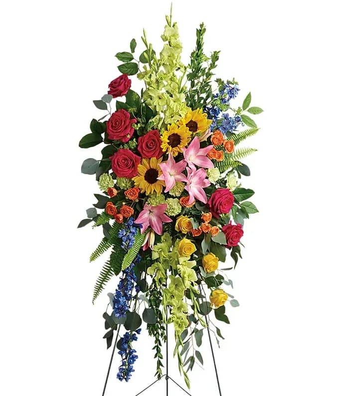 Tall standing spray of bright mixed flowers including roses, lilies, sunflowers, and delphinium on an easel