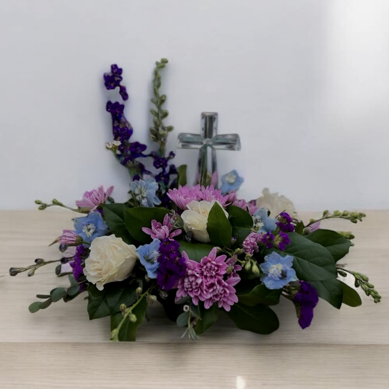 Low arrangement of white roses, lavender mums, and blue blooms with a glass cross in the center